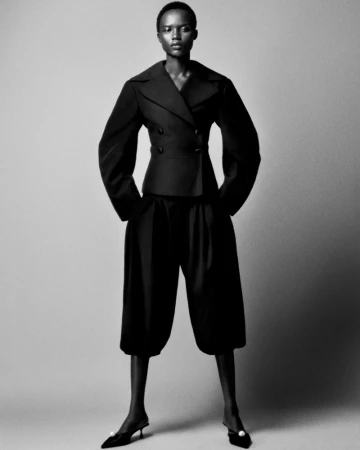 A black and white full shot of a slender Black woman in a dark suit jacket and wide-leg cropped pants against a grey studio backdrop.