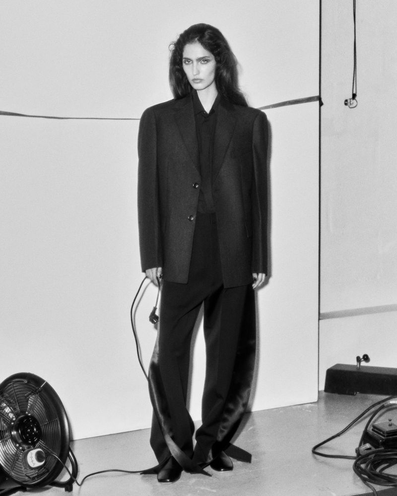 A slender woman in an oversized black blazer and trousers, standing against a white background in a studio, with fans and equipment.