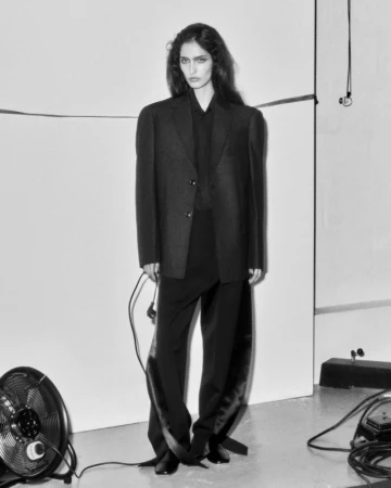 A slender woman in an oversized black blazer and trousers, standing against a white background in a studio, with fans and equipment.