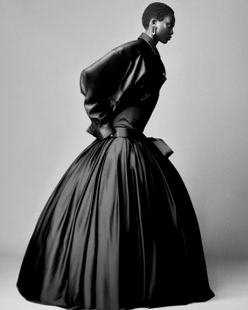 An adult female model poses in a photo studio wearing a black jacket and a voluminous black ballgown-style skirt against a neutral backdrop.