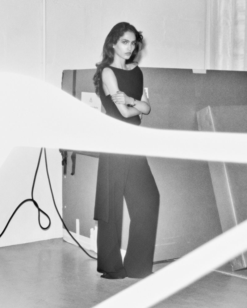 Elegant woman in black dress and wide-legged pants posing in an indoor studio with bright lighting, black and white photography.