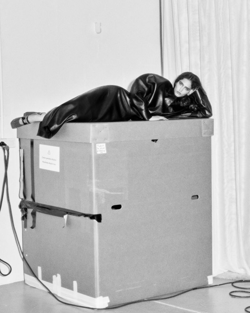 Woman in a black dress poses on a large box in a studio with a white backdrop, creating a monochromatic and minimalist fashion photo.