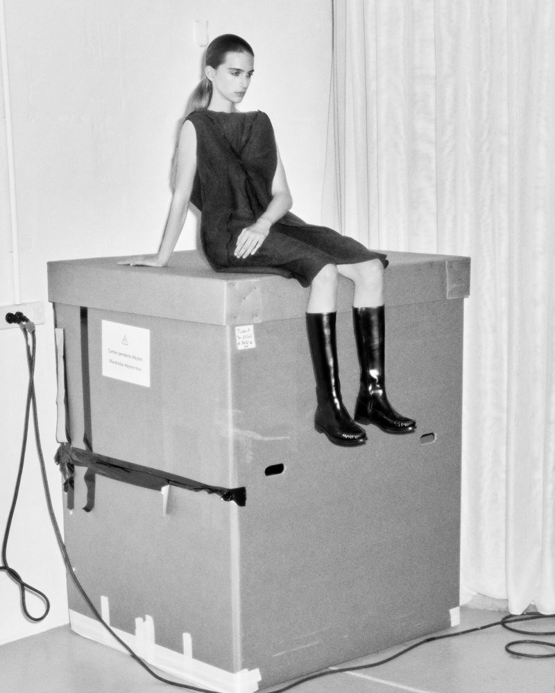 A fashion shot of a young adult woman posing on a large box, wearing a short dress and shiny knee-high boots, against a plain backdrop.