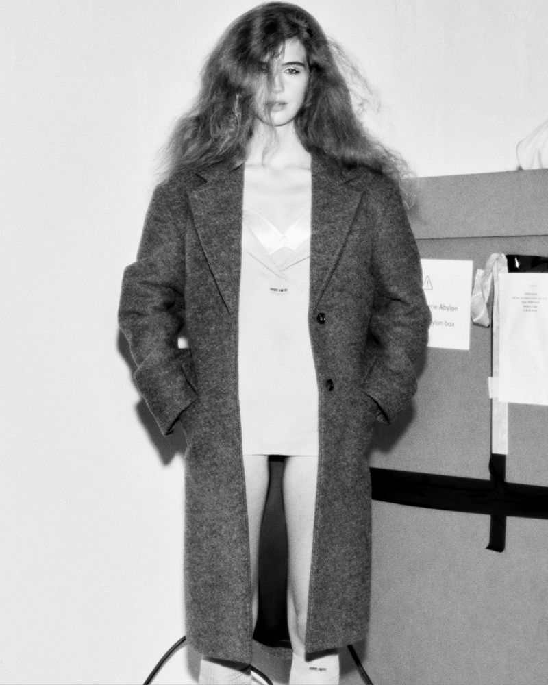 Black and white portrait of a woman with long wavy hair wearing a long coat in front of a backdrop.