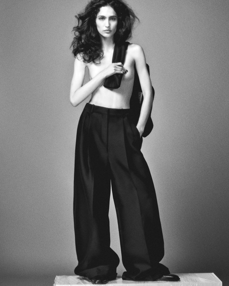 Black and white fashion shot of a topless woman in wide-leg trousers, carrying a jacket. Indoor studio setting with neutral backdrop.