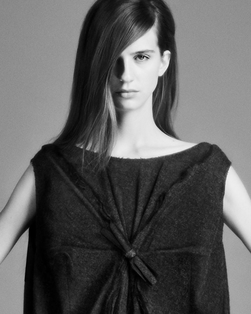 Black and white portrait of a woman wearing a dark dress. The image shows her upper body, arms down at sides, and long hair.