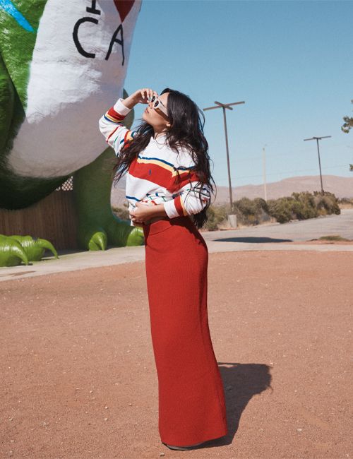 Woman in stylish red skirt and striped shirt poses next to the Cabazon Dinosaurs in a sunlit outdoor scene, creating a retro-inspired aesthetic.