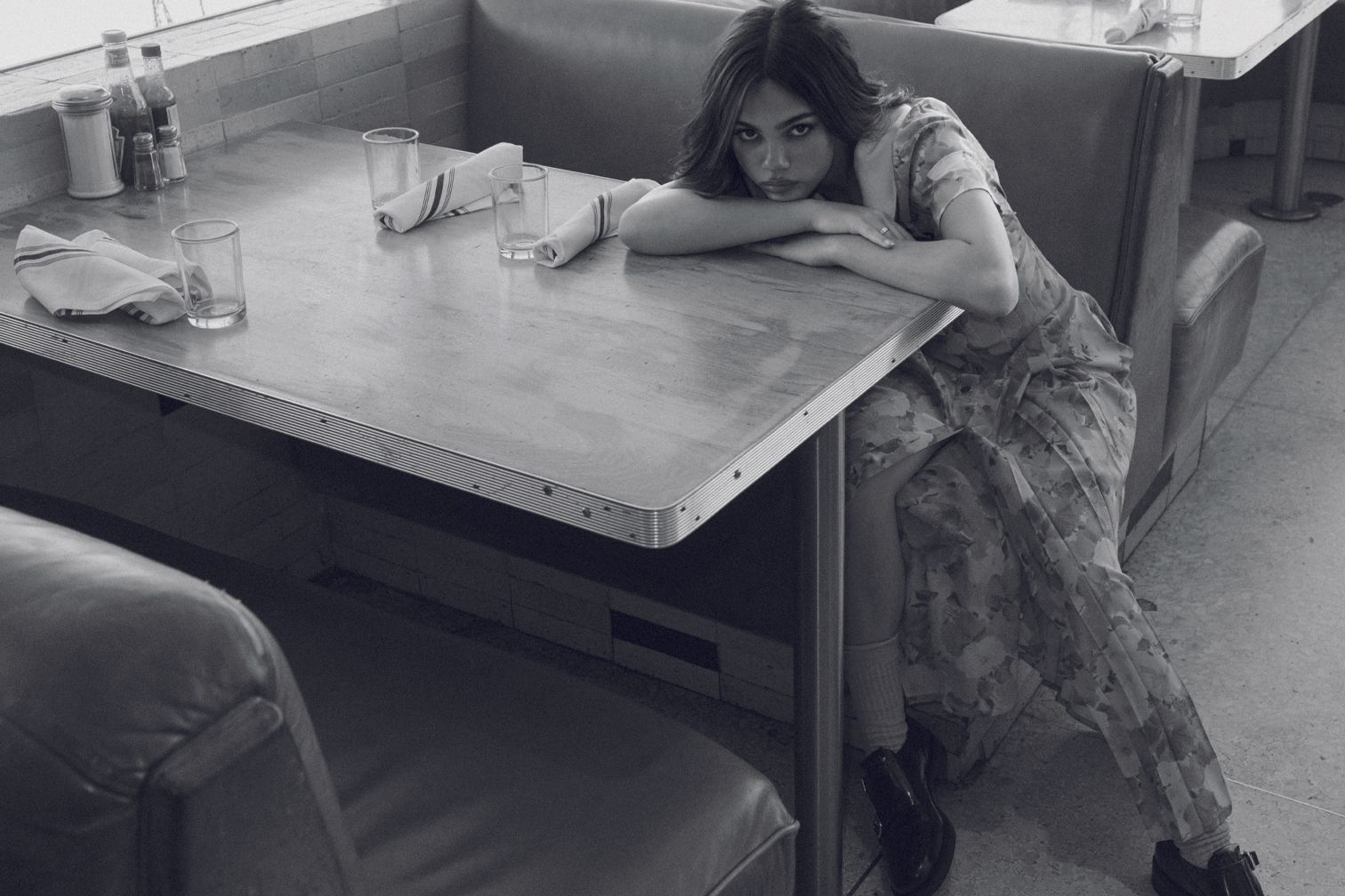 Woman in floral dress sits at retro diner table in shallow focus, with glasses, napkins, and condiments. Black and white photo.