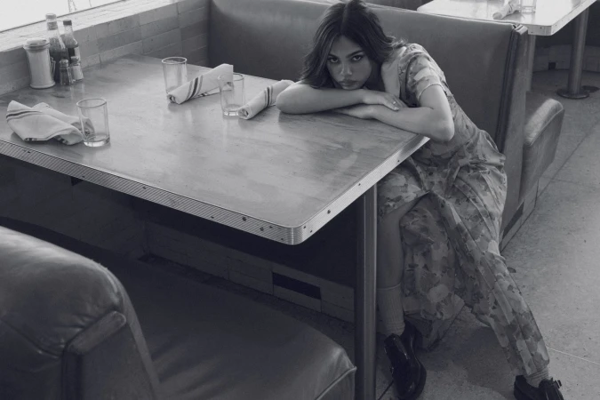 Woman in floral dress sits at retro diner table in shallow focus, with glasses, napkins, and condiments. Black and white photo.