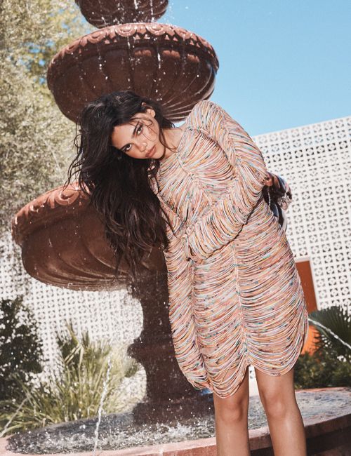 A woman in a patterned dress poses near a stone fountain with water flowing. She leans towards the camera in an outdoor setting.