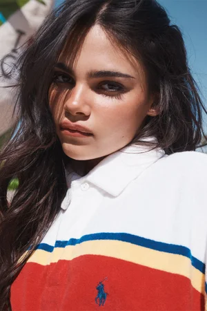 Close-up portrait of a dark-haired young woman wearing a white, red, and blue striped polo shirt under a bright sky.