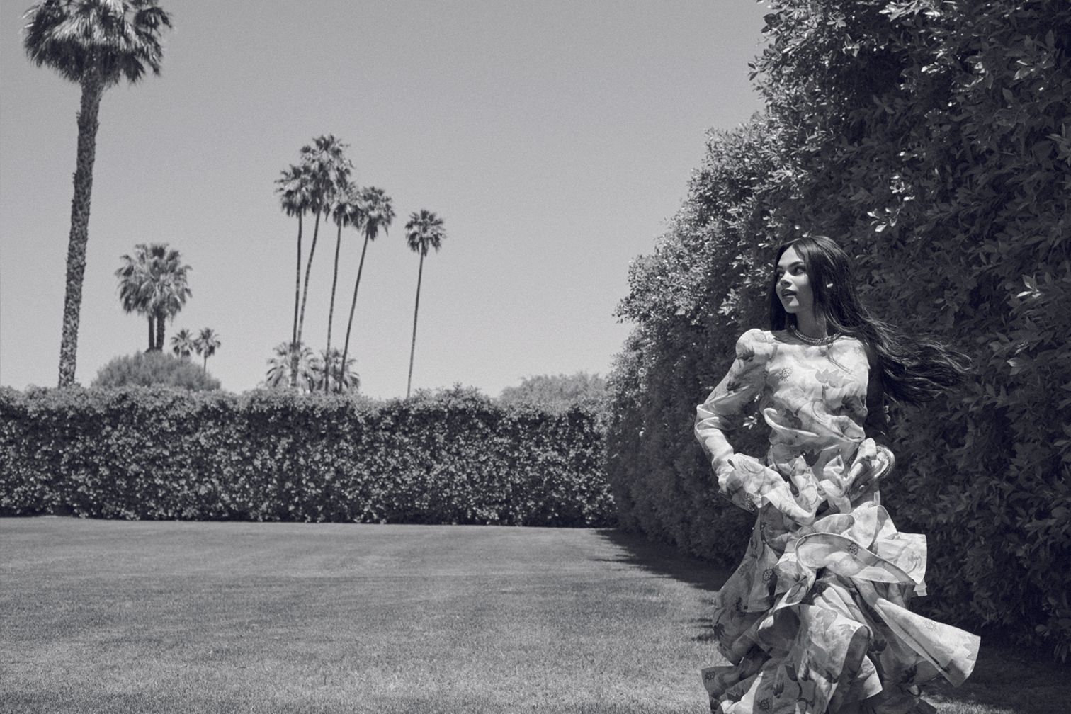 Elegant woman in floral dress stands on lawn, posing outdoors in a fashion shot. Palm trees and trimmed bushes in the background. Black and white.