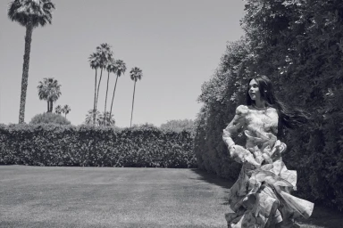 Black and white editorial portrait of a woman in a patterned ruffled dress running across a manicured lawn near tall palm trees.