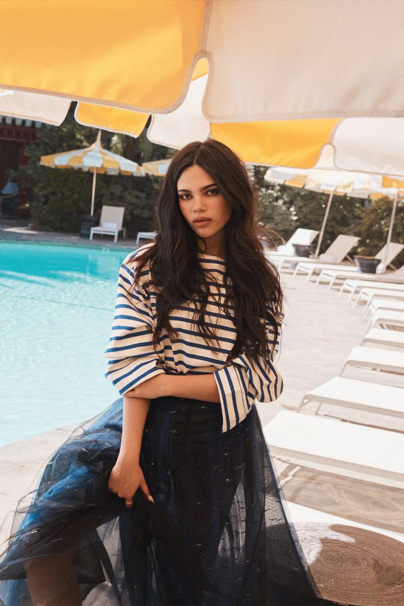 Woman in striped sweater and dark tulle skirt by a swimming pool under yellow umbrellas