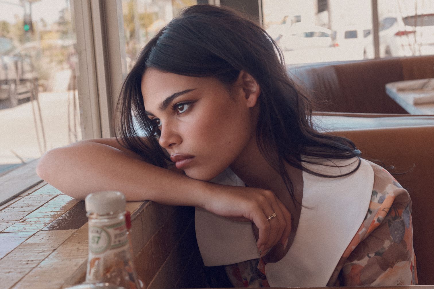 Brunette woman in floral dress looking out window with wistful expression in restaurant booth, capturing a vintage editorial aesthetic.