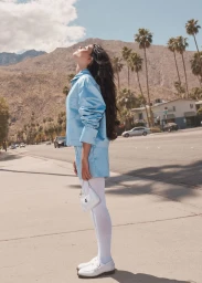 Woman with long dark hair wearing a light blue set and white tights standing on a sunny street with palm trees and mountains