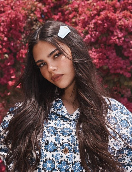 A young woman with long brown hair and a floral jacket poses in front of a vibrant pink flower bush for a portrait shot.
