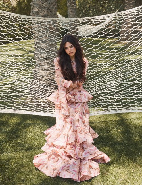 Brunette woman in a pink floral gown seated in front of a white net, surrounded by greenery in an outdoor setting, natural light.