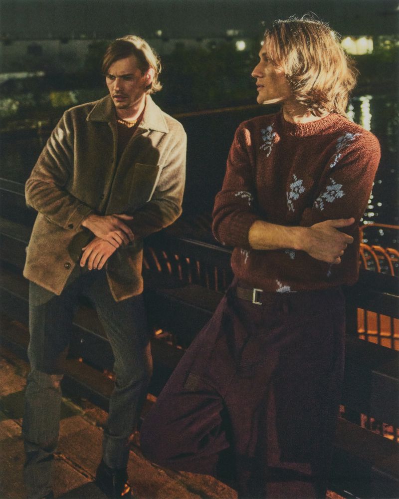 Two adult men pose on a bridge at night. One wears a brown jacket and jeans; the other a floral sweater and dark skirt.