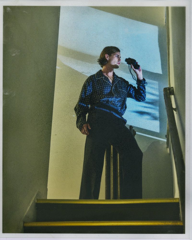 Adult male stands on a staircase in a patterned shirt, holding binoculars, looking toward a lit wall casting a shadow of window blinds.