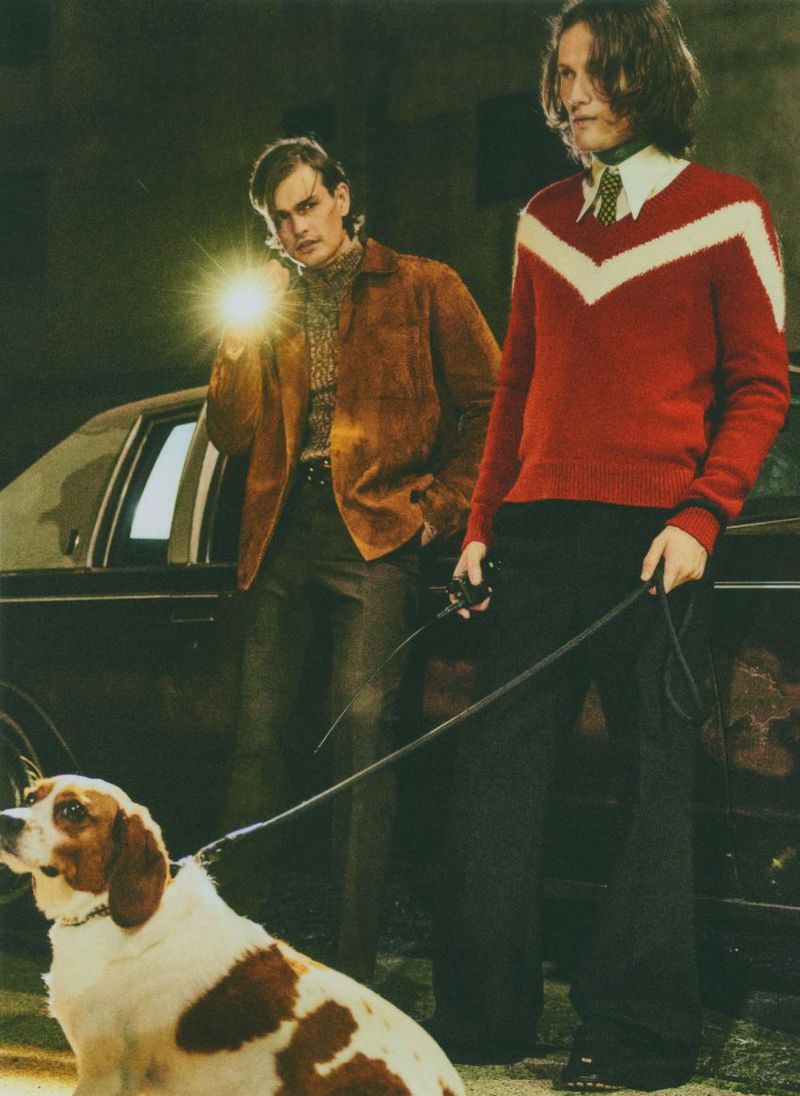 Two men stand with a dog near a car at night, one holding a flashlight, the other a leash, in a fashion or editorial style.