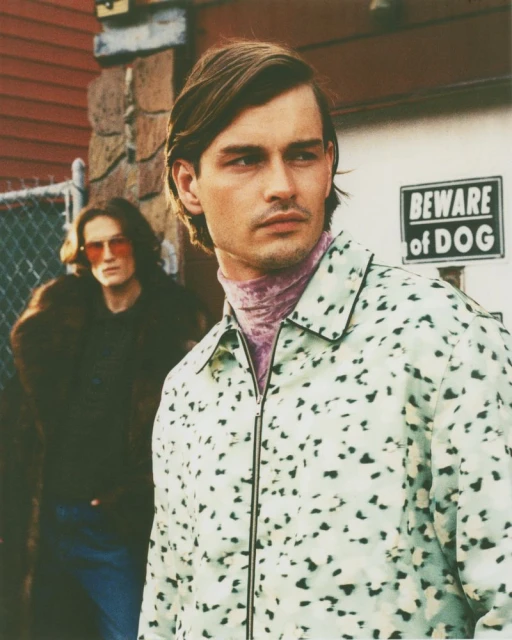 Two adult men in an outdoor urban environment. One man in the foreground in a patterned jacket; the other man wears sunglasses.