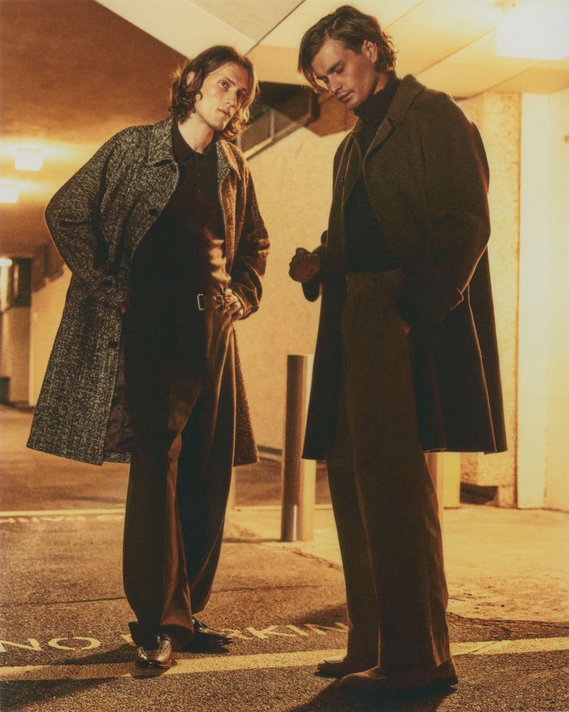 Two men standing in a parking garage wearing long coats and brown trousers. They are both looking down with a serious expression.