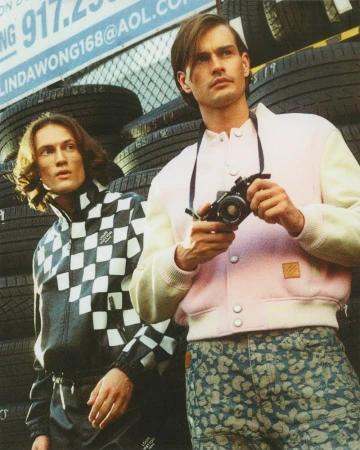 Two male fashion models pose outdoors in front of tires. One holds a vintage camera, wearing a pink bomber jacket, and patterned pants.