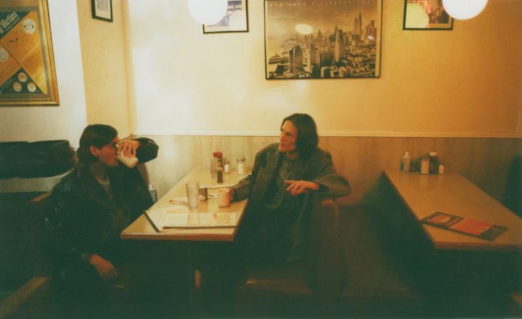 Two people seated at a booth table in a diner, one drinking, the other looking towards them in this warm-toned photograph.
