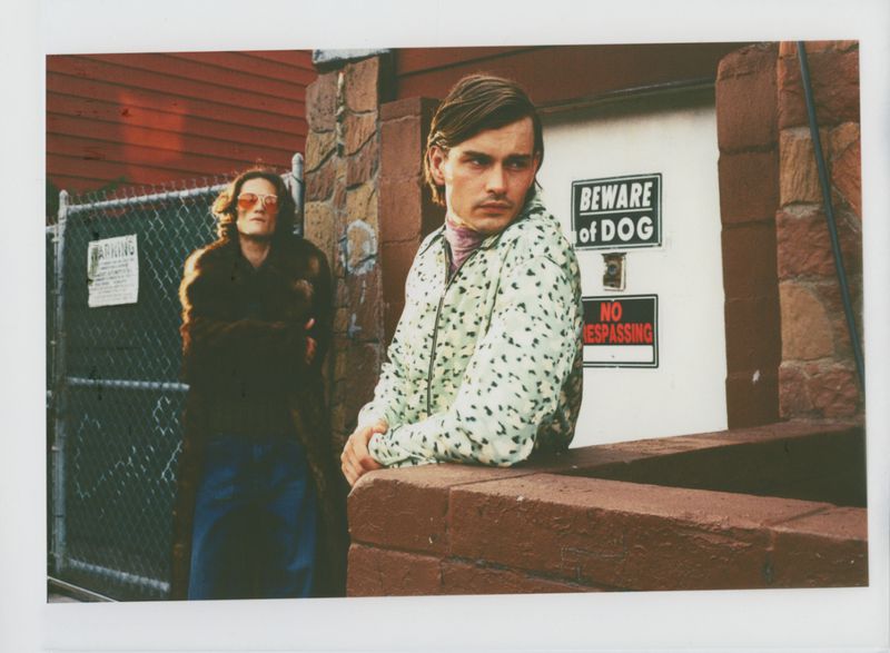 Two people pose outside a building; one in a dalmation-print jacket and the other wearing a brown fur coat and blue pants.
