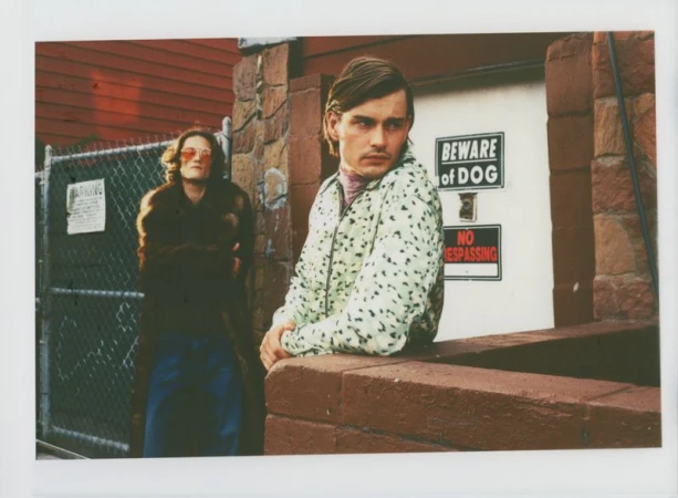 Two people pose outside a building; one in a dalmation-print jacket and the other wearing a brown fur coat and blue pants.