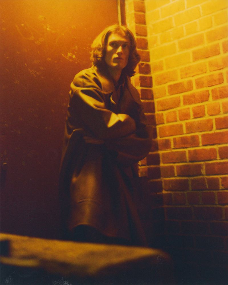Man in a coat stands against a brick wall under warm lighting. The scene has a gritty urban feel with a vintage aesthetic.