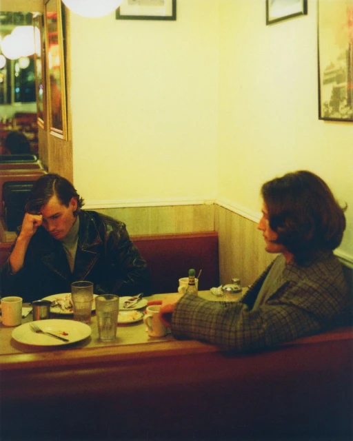 A man and a woman sit across from each other in a diner booth with plates of food, coffee, and condiments on the table between them.