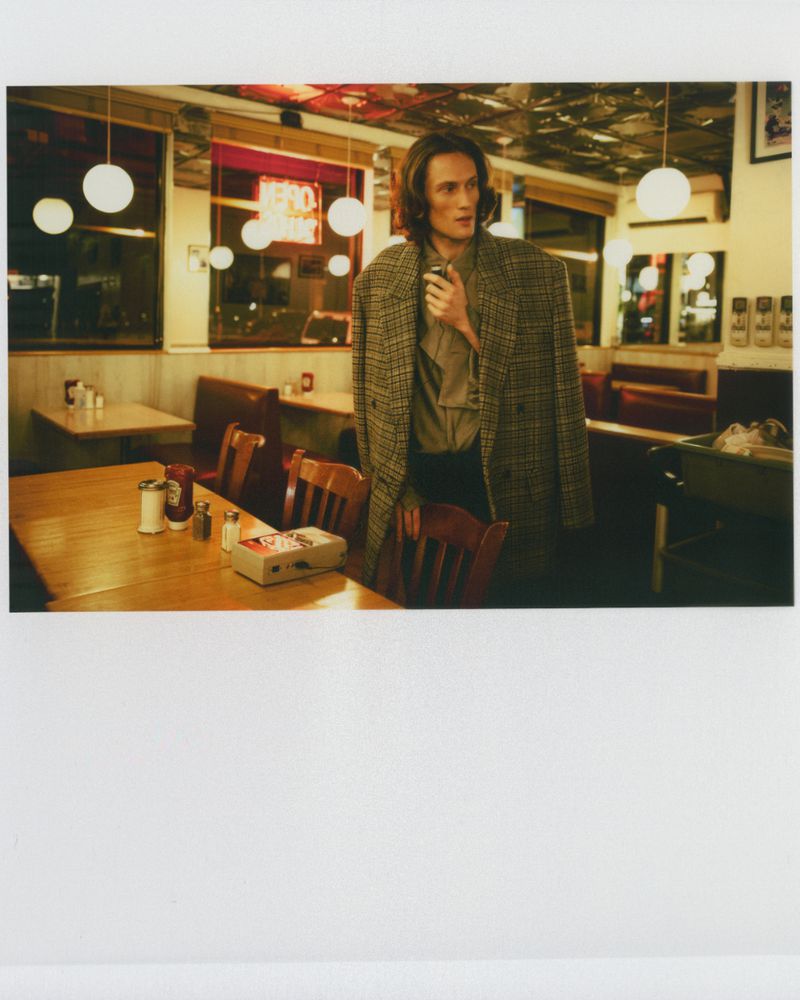 Man in a coat poses inside a diner near a wooden table with condiments and a vintage tape recorder, illuminated by warm lighting.