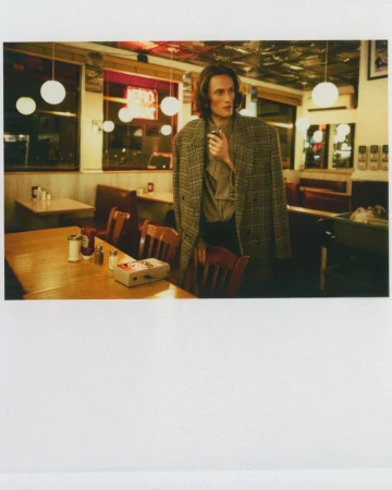 Man in a coat poses inside a diner near a wooden table with condiments and a vintage tape recorder, illuminated by warm lighting.