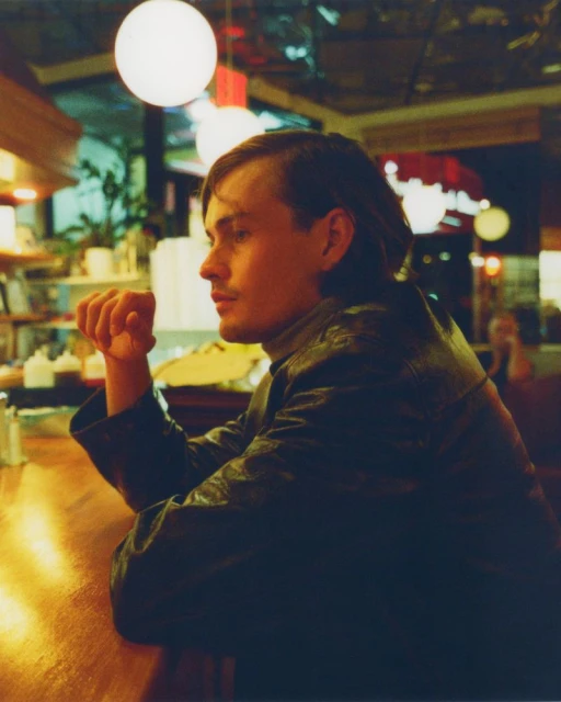 Adult male in a black jacket sits at a wooden bar indoors with spherical lights overhead, looking off to the side in a medium shot.