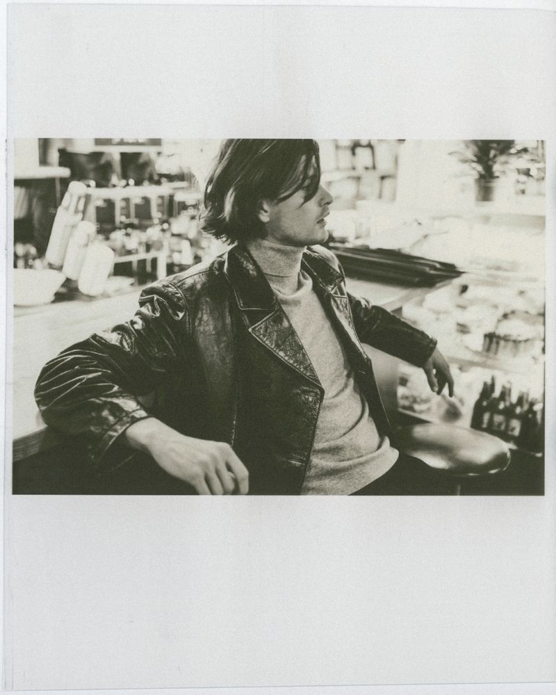 Adult male with long brown hair and leather jacket sits at counter inside with shelves and bottles visible in the blurred background.