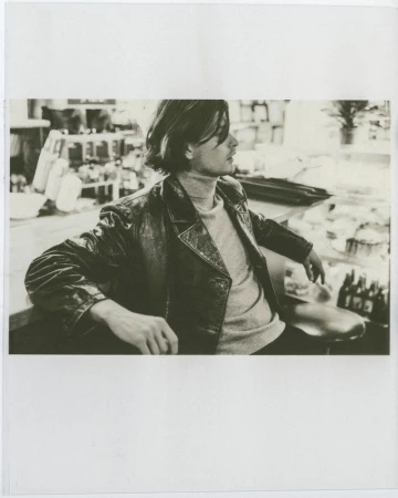 A man with dark hair wearing a leather jacket and turtleneck seated inside a cafe counter for GQ Portugal.