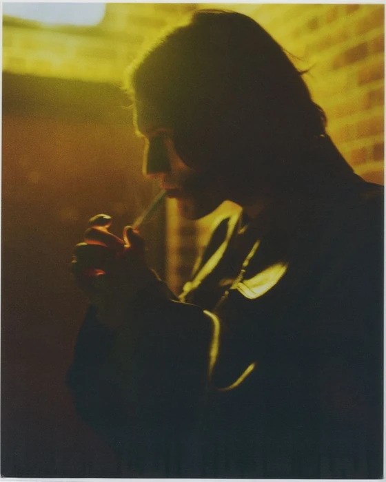 A man in dark jacket cupping his hands to light a cigarette under moody amber lighting against a brick wall.