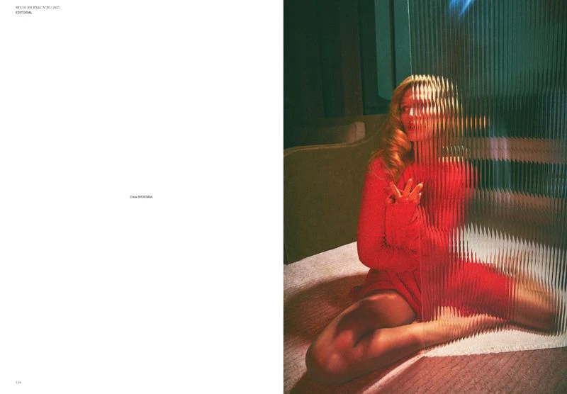 Woman in red dress sitting on bed behind textured glass panel, soft lighting, indoor setting, fashion, portrait orientation.