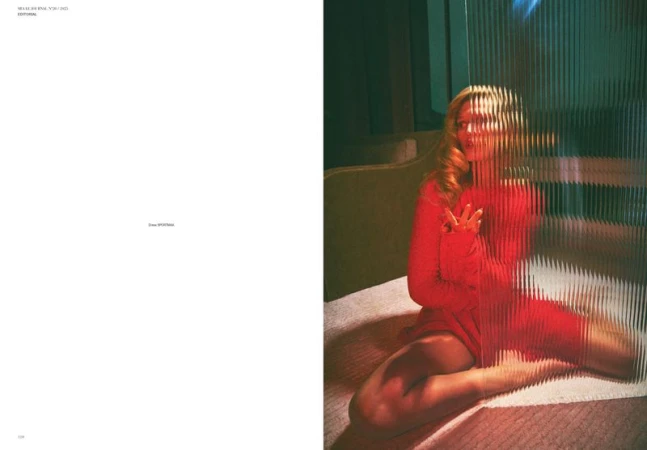 Woman in red dress sitting on bed behind textured glass panel, soft lighting, indoor setting, fashion, portrait orientation.