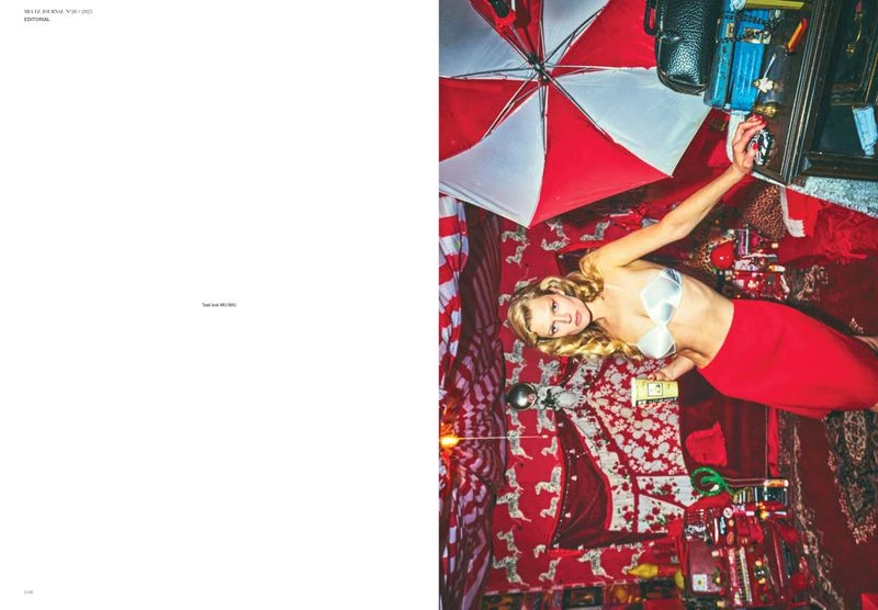 Editorial fashion shot of a blonde woman in a bra top and red skirt lying on a red patterned rug, surrounded by various objects indoors.