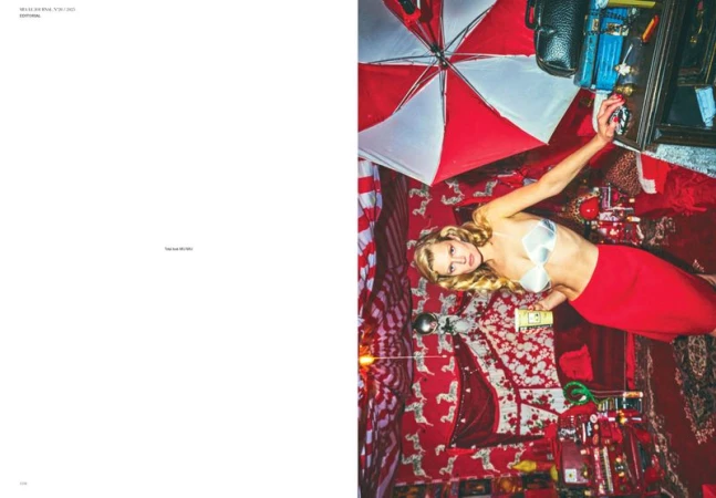 Editorial fashion shot of a blonde woman in a bra top and red skirt lying on a red patterned rug, surrounded by various objects indoors.