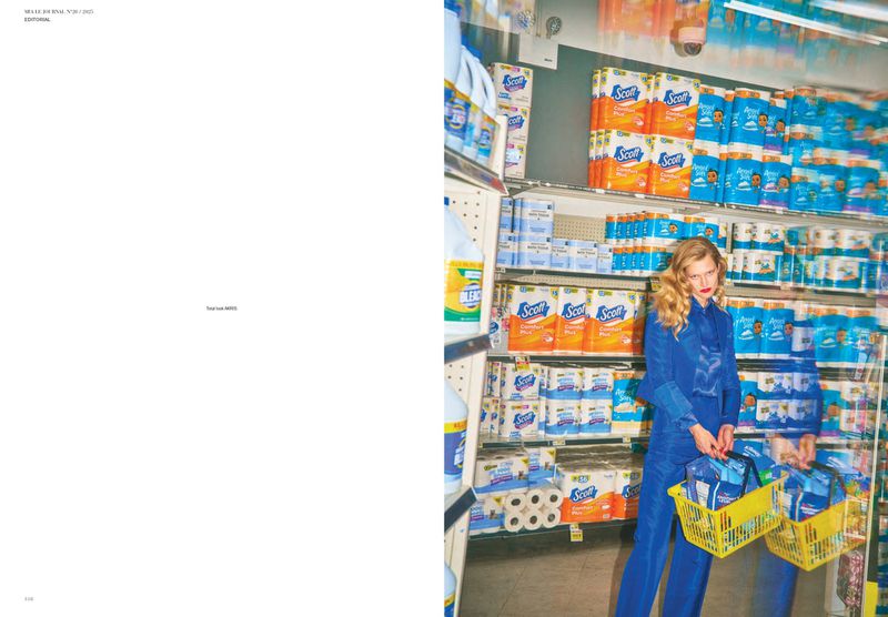 Woman in a blue suit stands in a supermarket aisle with shelves stocked with cleaning and paper products and carries a yellow basket.