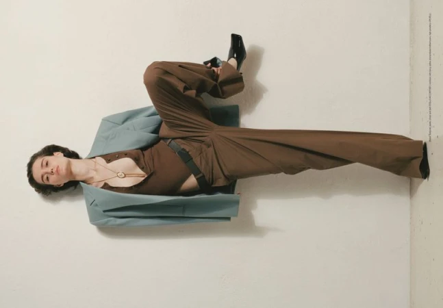 Model wearing brown trousers and a light blue jacket posed against a plain wall in an editorial fashion photograph.
