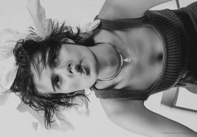 Monochrome portrait of a young adult female model with tousled hair, a neutral expression, and wearing a grey top and pendant necklace.