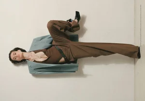 Model wearing brown trousers and a light blue jacket posed against a plain wall in an editorial fashion photograph.