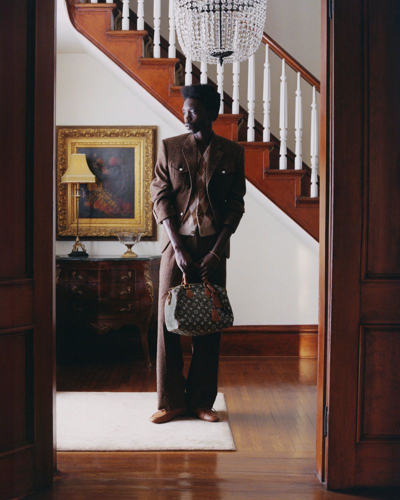 An African-American man in a brown suit poses indoors, holding a monogrammed handbag. An ornate staircase and chandelier are in the background.