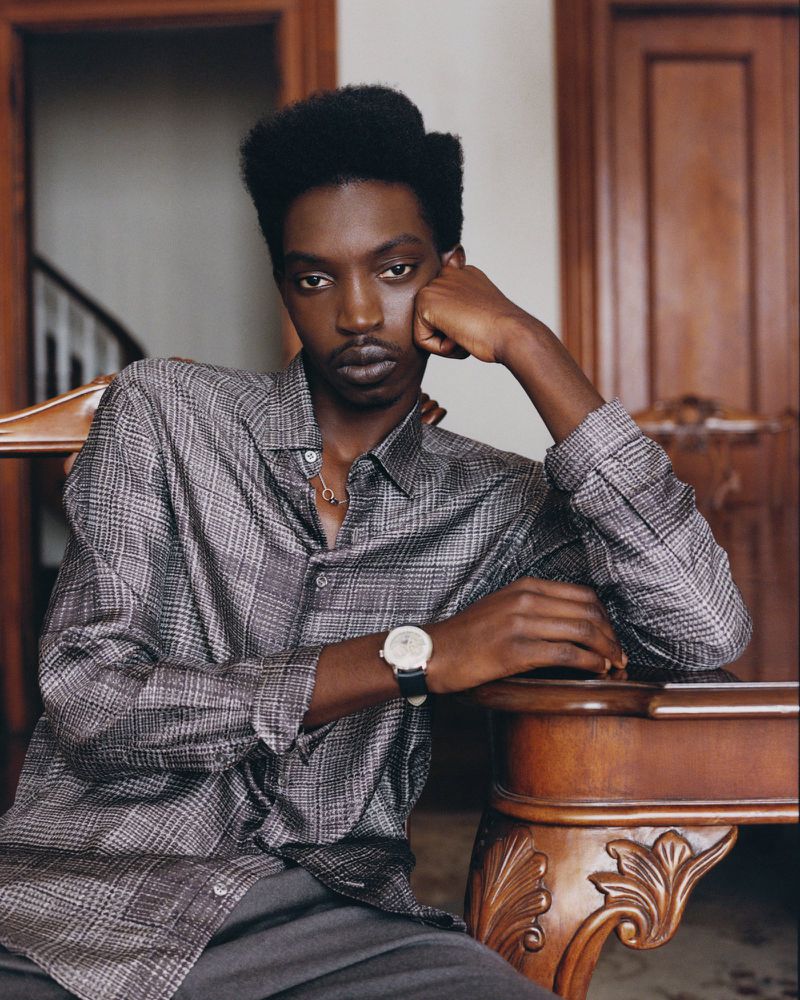 Model Lamine Seck wearing a grey plaid shirt, leaning on a wooden table in an interior editorial portrait.
