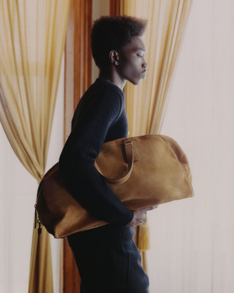 An African-American model carries a tan duffel bag in a studio with gold curtains, showcasing fashion and travel themes.
